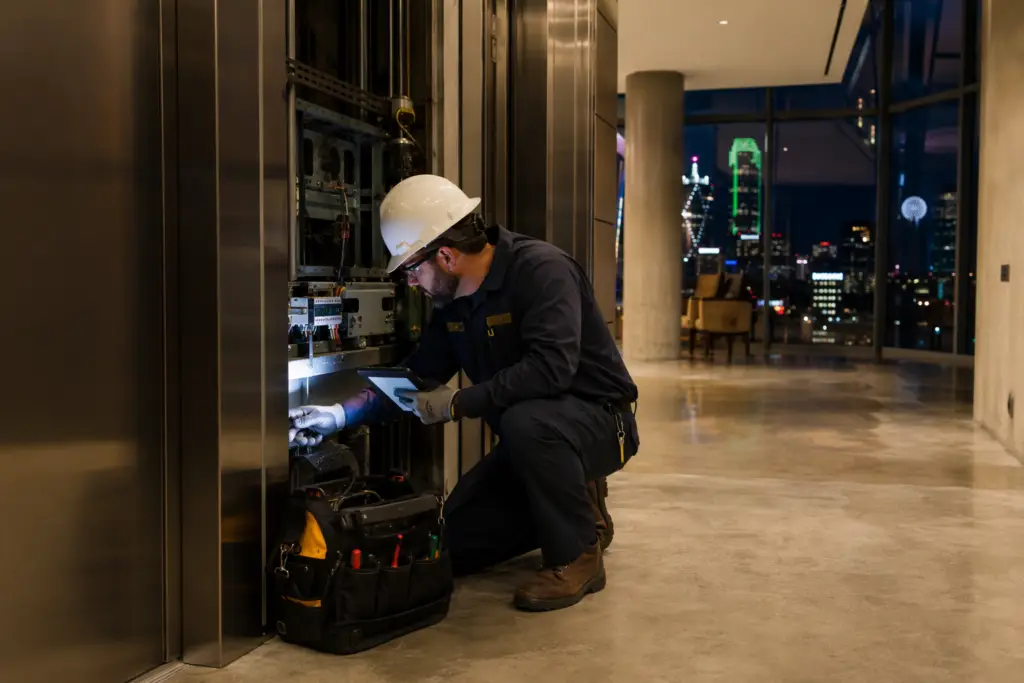 Certified elevator technician performing emergency elevator repair in a Dallas TX high-rise lobby at night, diagnostic tablet in hand beside open elevator panel.