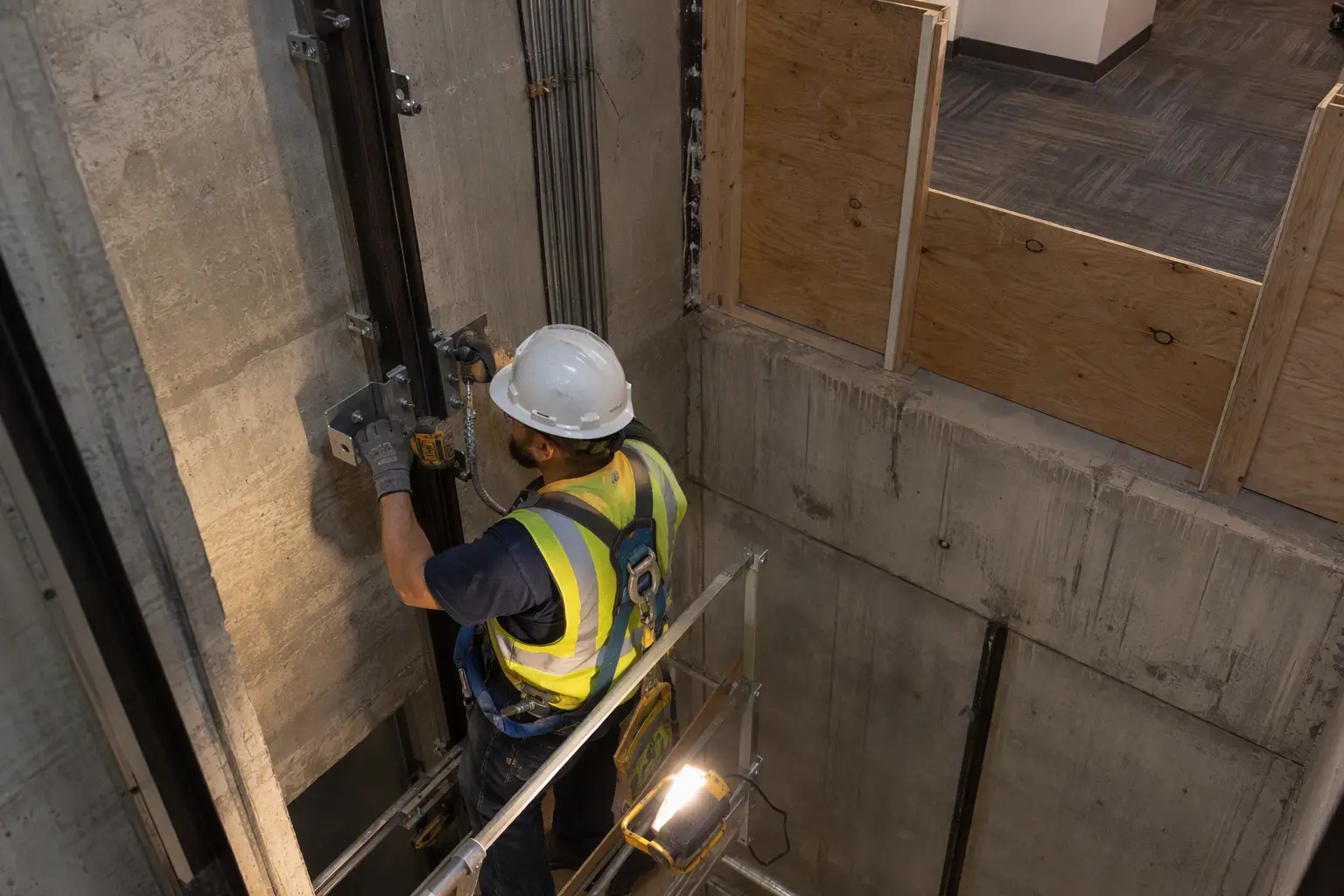 Elevator technician installs guide rail brackets inside a hoistway pit during phased new elevator construction in an occupied Houston building, with OSHA-compliant barriers separating the work zone fr
